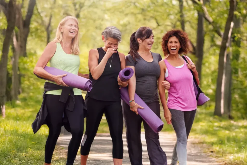 Four active senior women laughing and walking outdoors with yoga mats, enjoying a wellness activity in a green park.